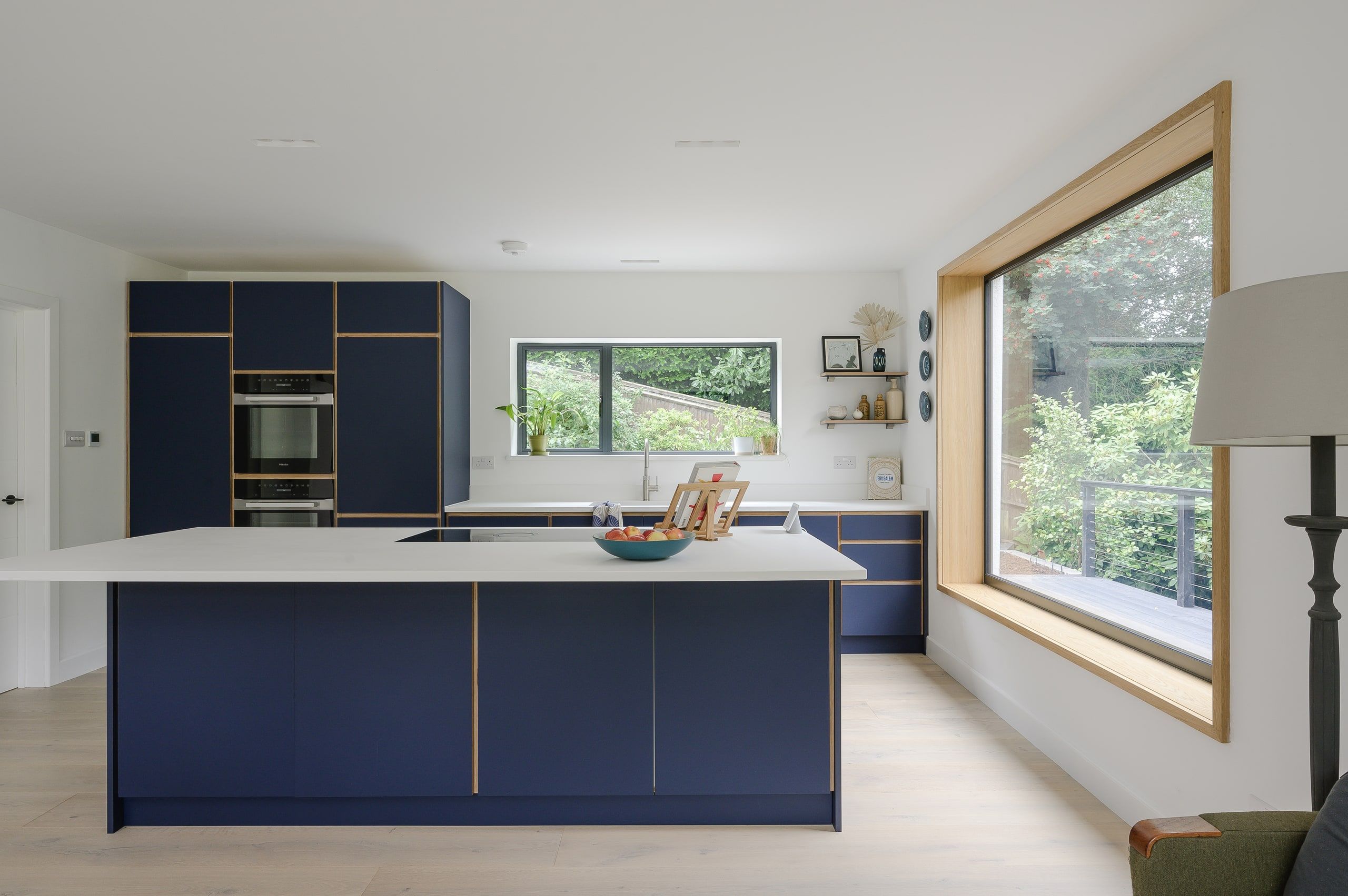 Open kitchen with a navy island, white worktops and an oak-framed picture window, 1960s house, Haslemere.