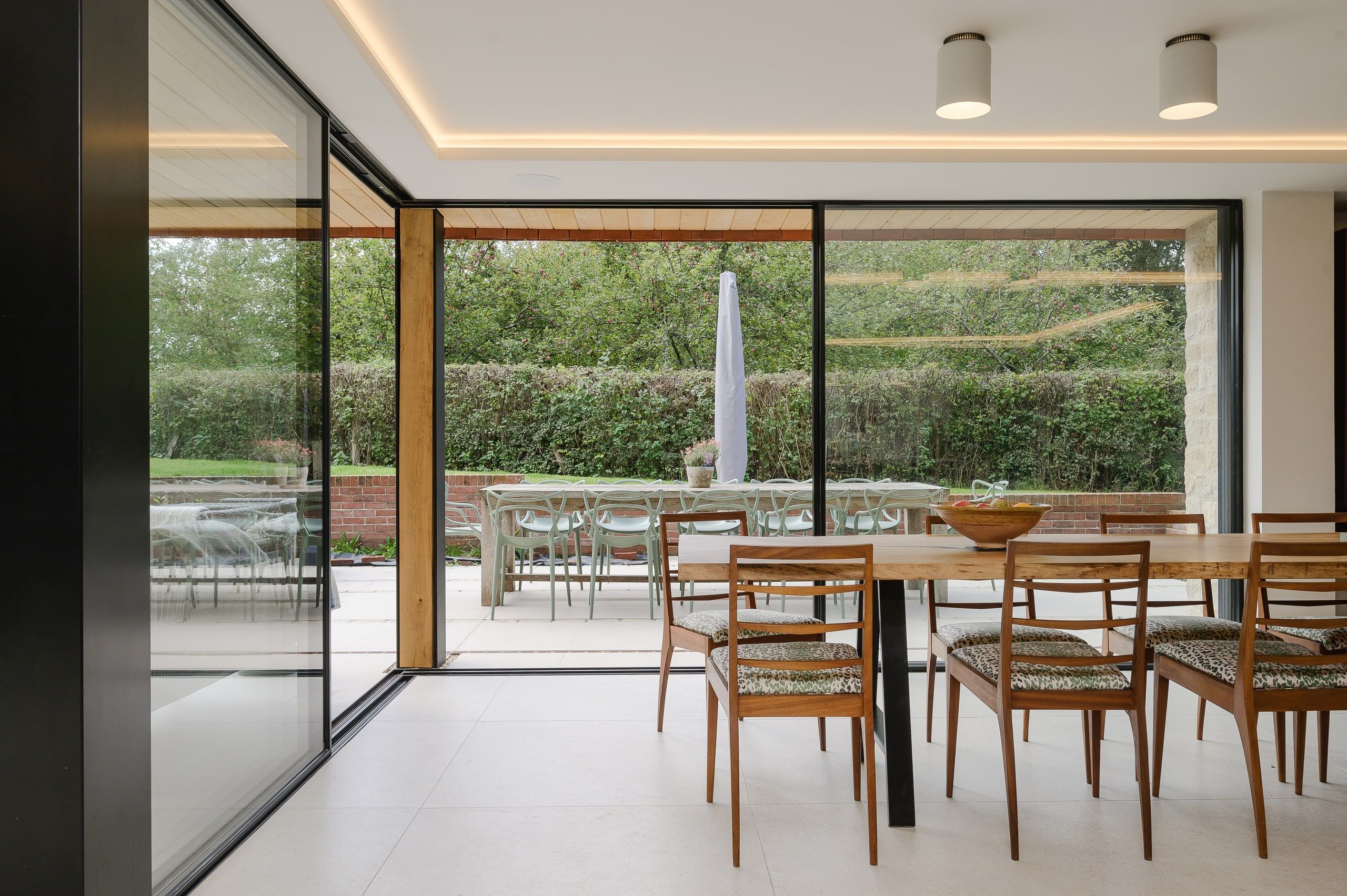 Dining table beside sliding glass doors opening to the terrace and clipped hedge, country house, Lodsworth.