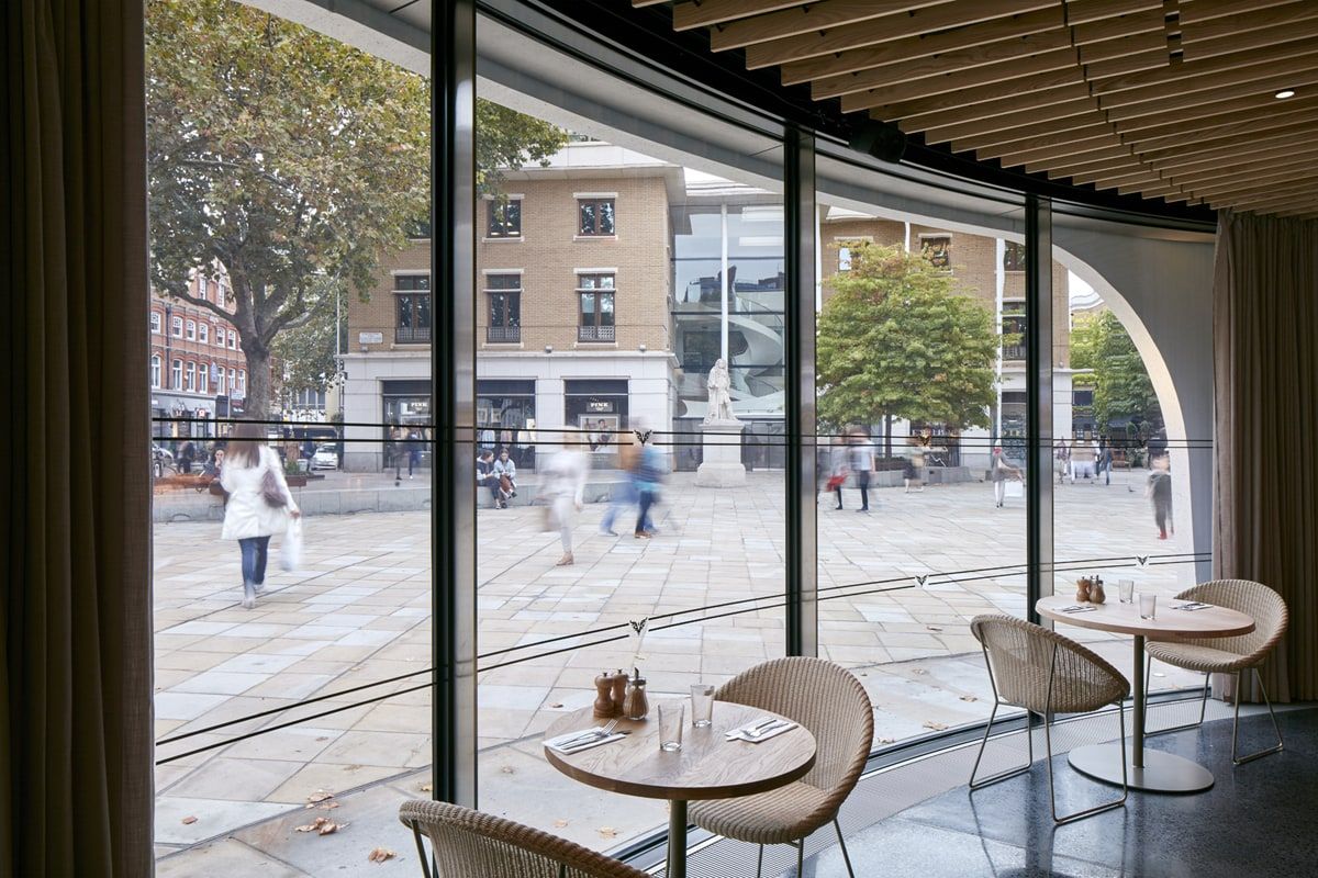Tables set along the curved glazing, with shoppers passing outside, restaurant pavilion, Duke of York Square, London.