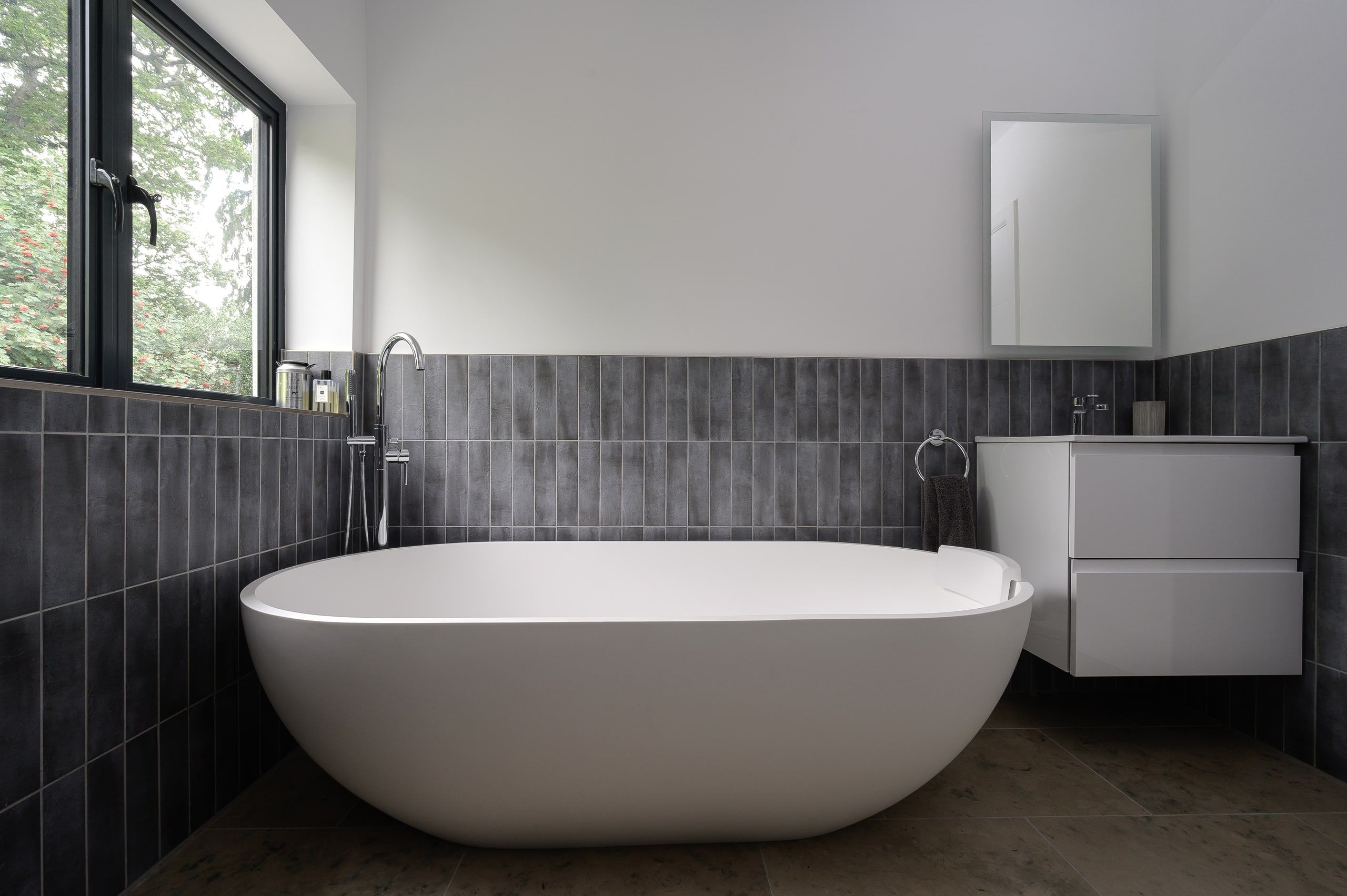 Freestanding oval bath against charcoal tiling below a black-framed window, 1960s house, Haslemere.
