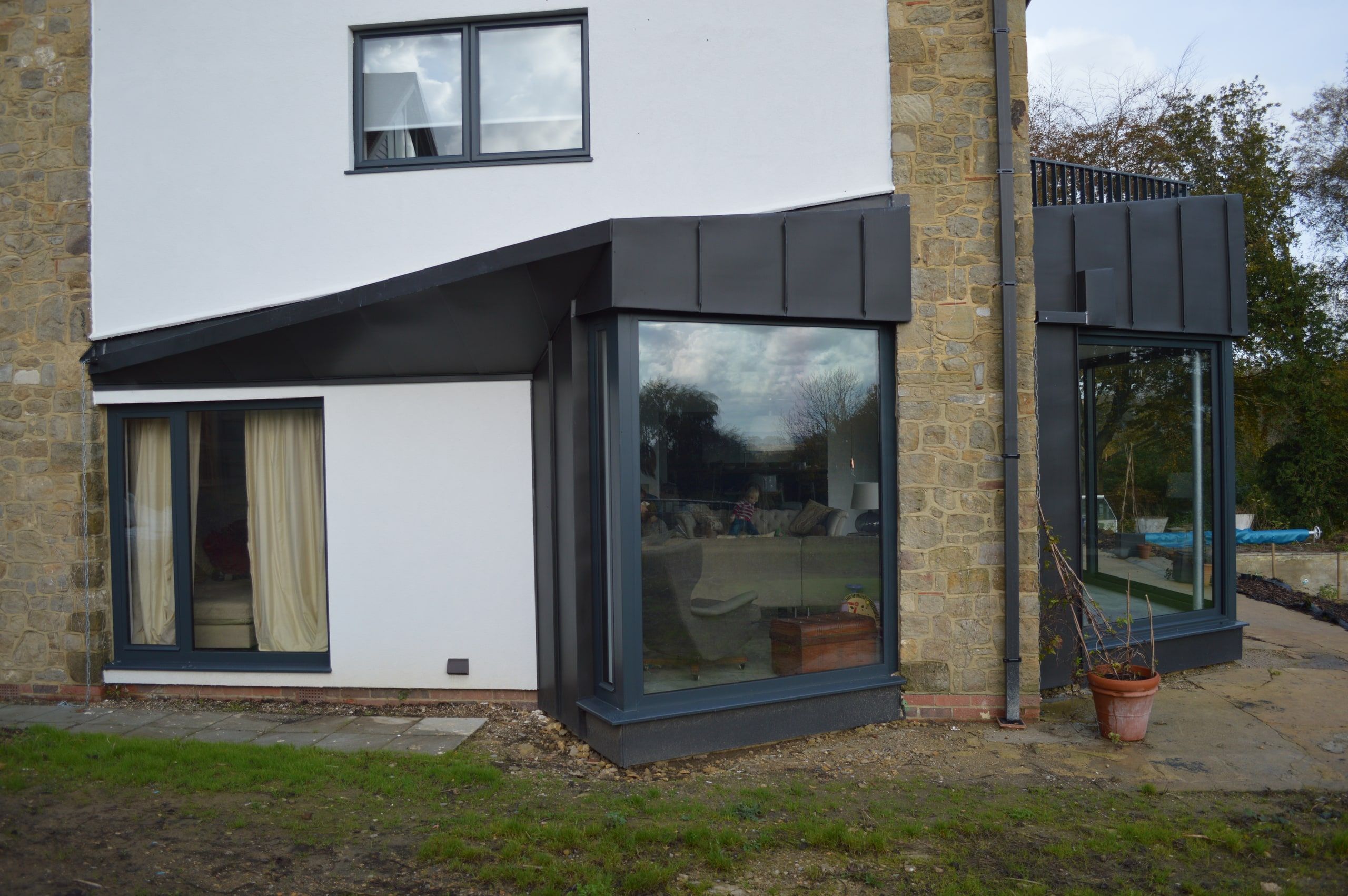 Two dark zinc-clad bay extensions on the rendered and stone garden elevation, 1960s house, Lodsworth.